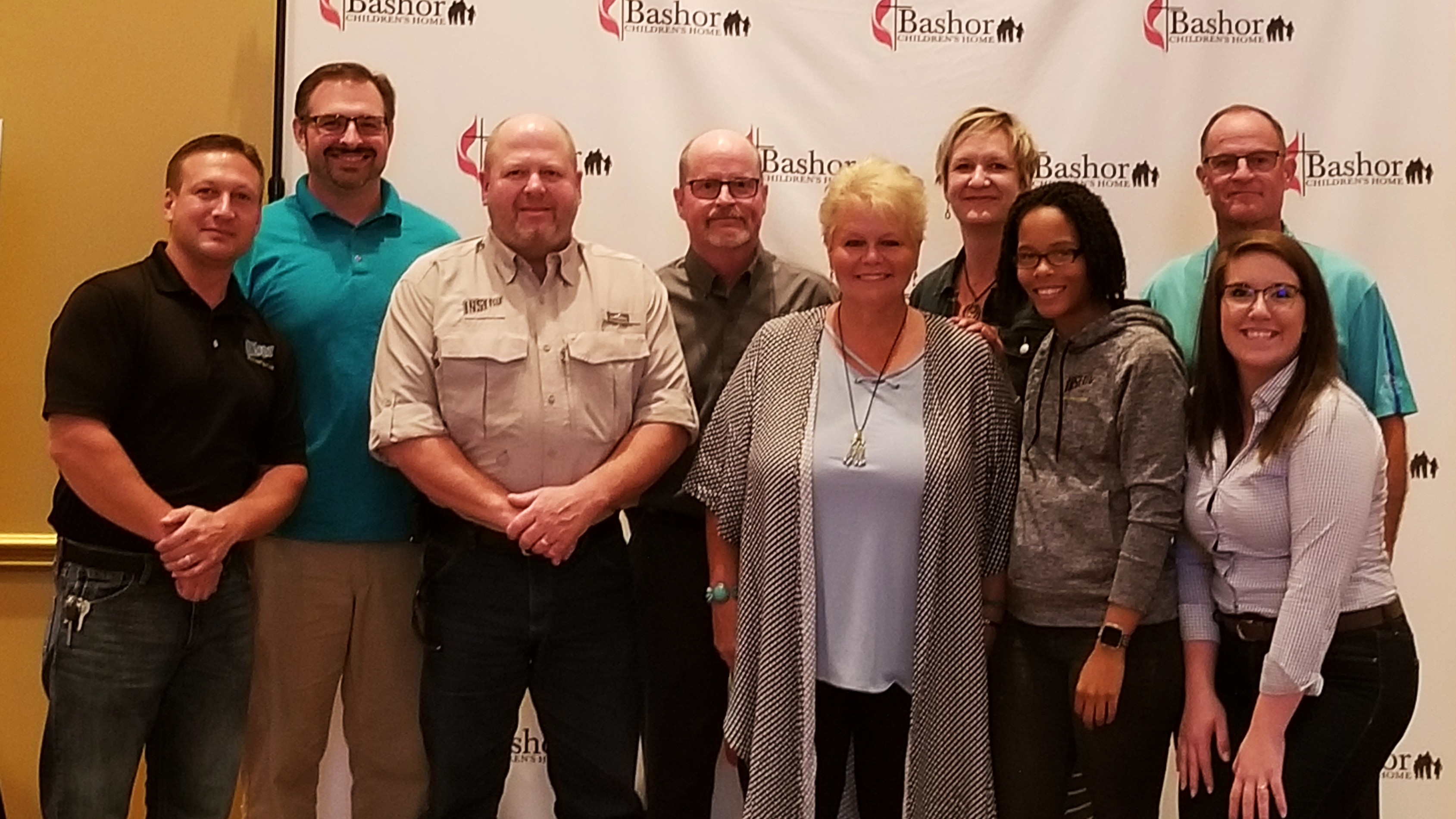 A group of nine INSERV employees stands together for a photo in front of a Bashor backdrop. The individuals include three men in the front, three women in the back, and a mix of casual and professional attire, with varying hairstyles and ages.