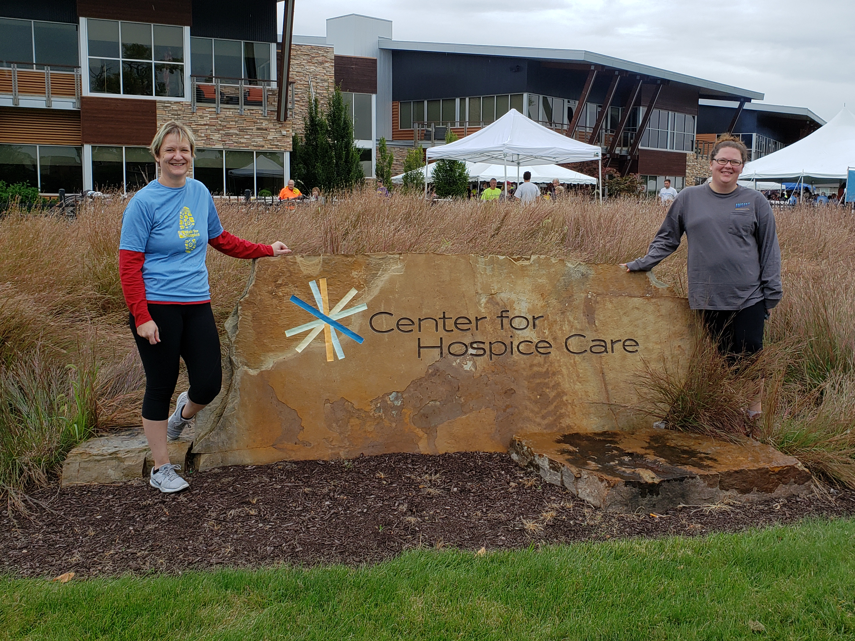 Two women stand beside a large stone sign displaying the text 'Center for Hospice Care.' The sign is surrounded by tall grass and greenery, with a modern building and white tents visible in the background under a cloudy sky.