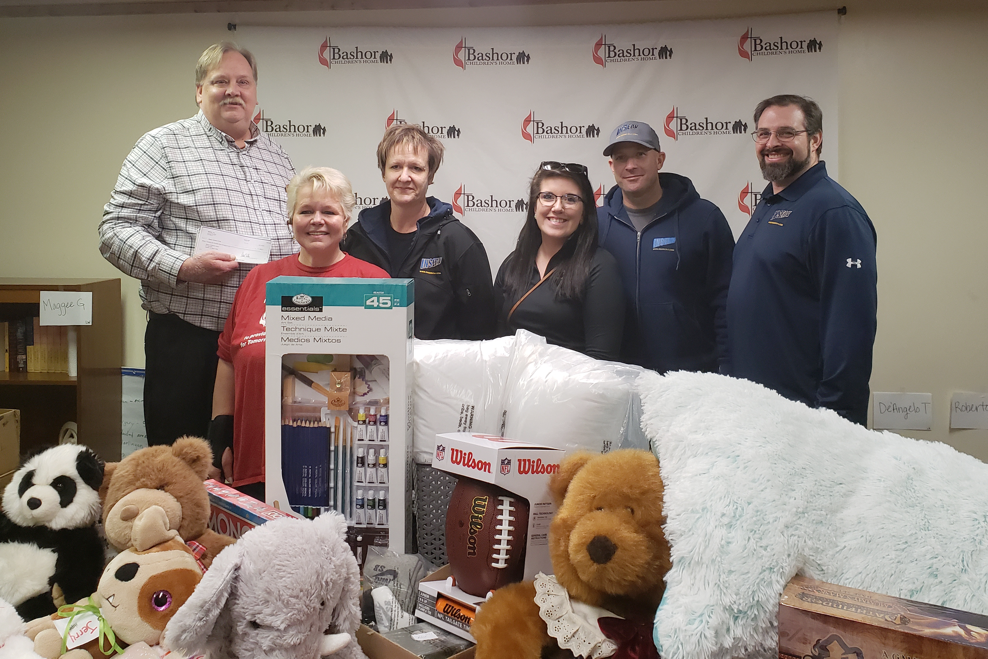A group of five individuals poses together in front of a banner featuring the logo 'Bashor.' They are surrounded by various items, including stuffed animals, bedding, and a package of supplies. One person holds a check in their hand.