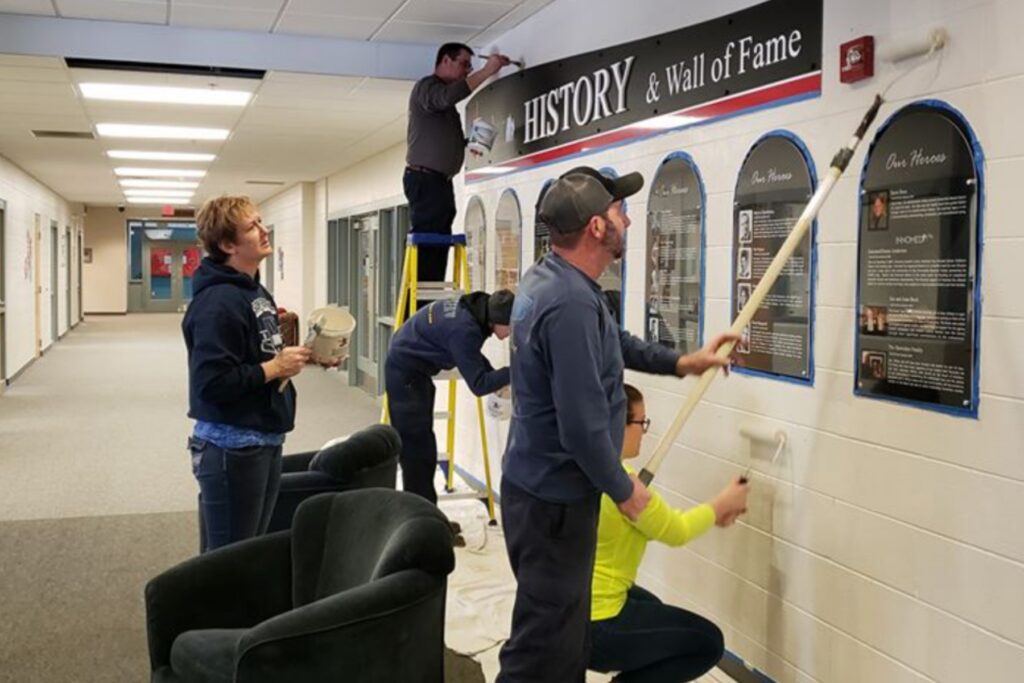 Five INSERV employees are working together to paint a wall in a hallway at Bashor Children's Home. One person is using a paint roller while standing on a ladder, another person is seated in a green chair, and the remaining three are holding paint supplies or preparing for painting. A banner above reads "HISTORY & Wall of Fame."