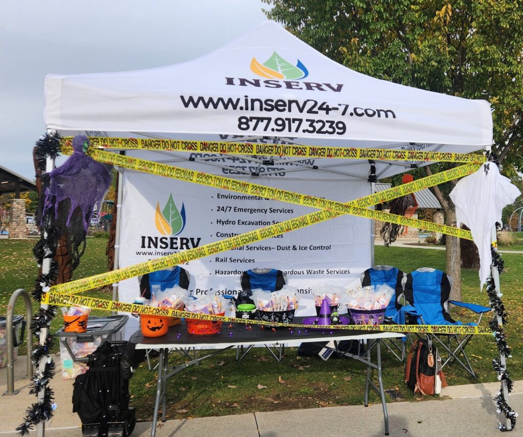 A decorated INSERV promotional tent with yellow caution tape across the front, set up on a sidewalk near a grassy area with trees.