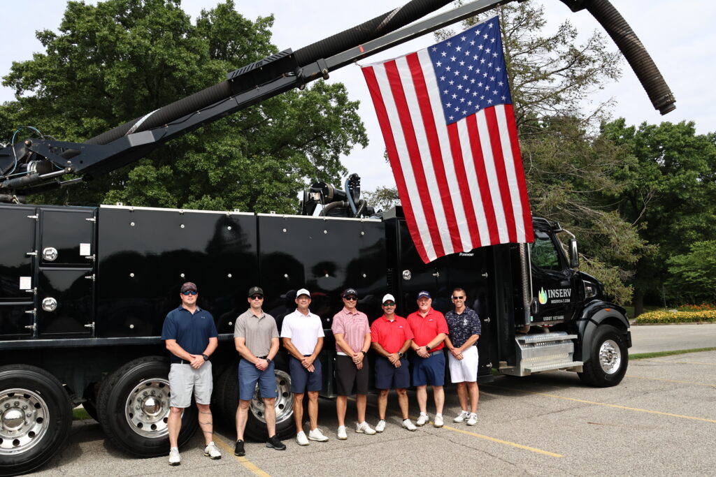 A group of eight men standing in front of a large black truck with a vacuum attachment. The truck features an American flag hanging from the top. The men are dressed in casual golf attire, including polo shirts and shorts, positioned in a row.
