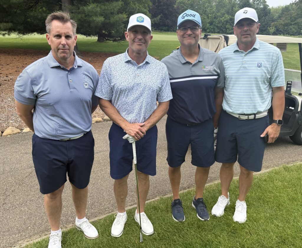 Four men pose on a golf course, standing on a paved path. The man on the left wears a gray polo and dark shorts, the second man wears a patterned white shirt and holds a golf club, the third man in a gray shirt smiles, and the fourth wears a light blue polo.