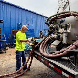 Environmental Services An industrial operator handling a heavy transfer hose at the back of an INSERV vacuum truck parked outside a facility.