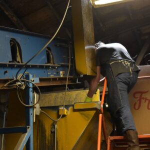 Environmental Services A worker in an apron and boots standing on a rolling ladder to inspect or service heavy sorting machinery inside a dimly lit facility.