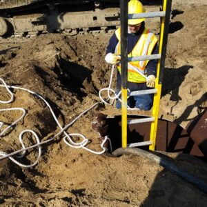 Environmental Services A utility worker wearing safety gear manages a rope near a ladder leading into an underground excavation hole.
