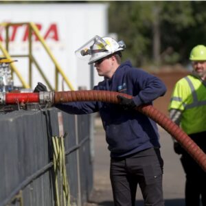 Environmental Services An industrial technician wearing a hard hat with a face shield manages a large transfer hose connected to a storage containment unit.