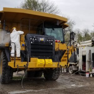Environmental Services An industrial technician in a hazmat suit uses a high-pressure power washer to clean a massive yellow CAT mining dump truck on a muddy site.