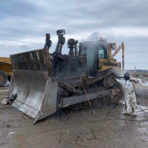Environmental Services An environmental technician in a white protective suit uses a high-pressure steam power washer to clean a large, muddy CAT bulldozer.