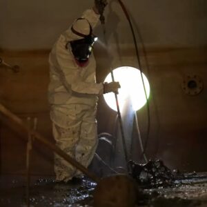 Industrial Services An industrial technician in full protective gear and a headlamp operating a pressure wand inside a dark, confined storage tank.