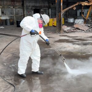 Industrial Services An industrial technician in a hazmat suit and respirator uses a high-pressure steam power washer on a warehouse floor.