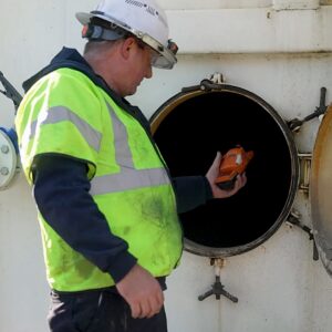 Industrial Services An industrial safety technician holding a gas detector device inside the open hatch of a white storage tank.