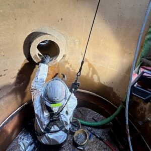 Industrial Services An industrial safety technician holding a gas detector device inside the open hatch of a white storage tank.