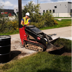 Emergency Spill Services A worker in a yellow safety vest and hard hat operates a small remote-controlled excavator on a grassy area beside two black barrels. In the background, a gray building and trees are visible.