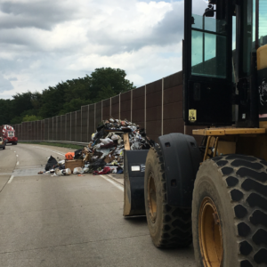 Emergency Spill Services Heavy construction loader positioned on a highway shoulder next to a large pile of spilled debris along a sound wall.