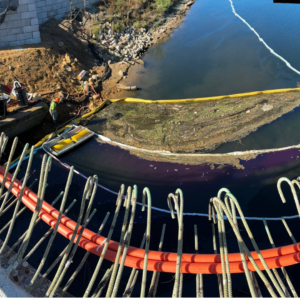Emergency Spill Services High-angle view of an environmental response site with containment booms and a skimmer deployed along a waterway near a construction zone.