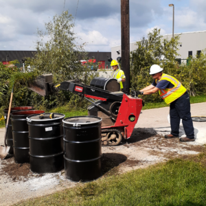 Emergency Spill Services Environmental response workers operating a mini track loader to deposit soil and vegetation into steel disposal drums.