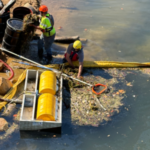 Emergency Spill Services Environmental cleanup workers in protective gear using an oil skimmer and hand tools to remove debris and contaminants from a body of water.