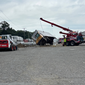 Emergency Spill Services Heavy tow truck using a crane boom to lift and upright an overturned box truck at an industrial worksite.