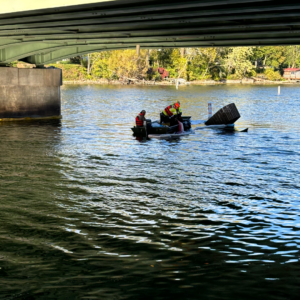 Emergency Spill Services Emergency response crew in a small boat deploying containment booms near a partially submerged object under a bridge.