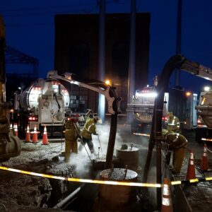 Hydro Excavation Services Nighttime hydro excavation operation with workers using pressurized lances and vacuum boom hoses around steam-emitting underground utility pipes.