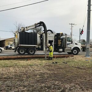 Hydro Excavation Services A utility worker in high-visibility gear operating a vacuum truck suction hose near a wooden utility pole beside a road.