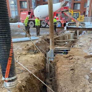 Hydro Excavation Services Construction workers standing near a deep excavation trench with concrete mixing equipment and a suction hose setup.