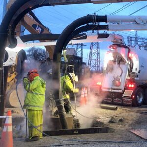 Hydro Excavation Services Dusk hydro excavation work with crew using steam pressure wands and vacuum hoses near heavy equipment and electrical infrastructure.