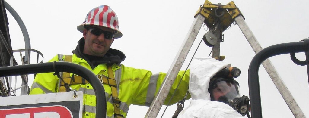A worker in a yellow safety jacket and hard hat stands beside a person in a white protective suit on top of a rail car. A sign attached to the rail car cautions about confined spaces and provides emergency contact information.