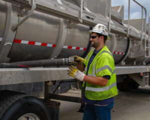 Careers A man wearing a safety helmet and a yellow reflective vest stands beside a silver tanker truck. He holds a black hose attached to the truck, which features red reflective stripes. The background shows a complex of industrial structures.
