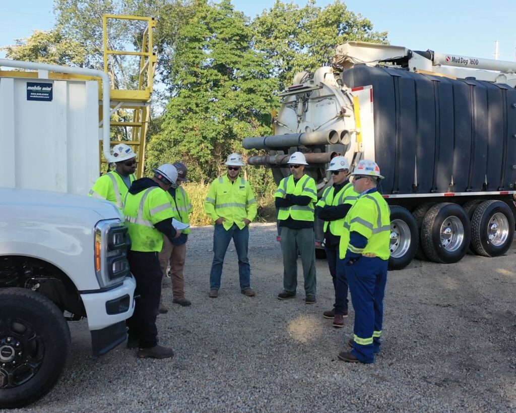 A group of eight workers in safety gear and helmets are gathered near a large truck and a white pickup truck at a work site. The background features trees and a storage container, with equipment visible in the scene.