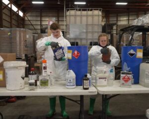 Careers Two technicians in protective suits inspect and sort various household and chemical waste containers at folding tables inside a facility.
