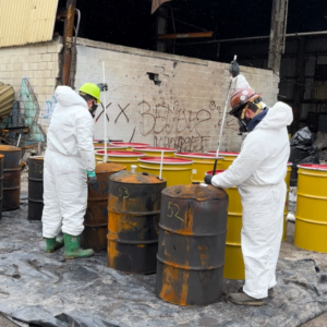 Two environmental response technicians in hazmat suits taking liquid samples from numbered chemical storage drums outdoors.