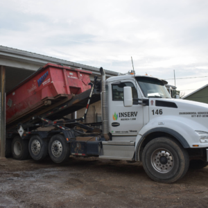 A white INSERV roll-off truck unloading or loading a red waste dumpster container outside a facility building.