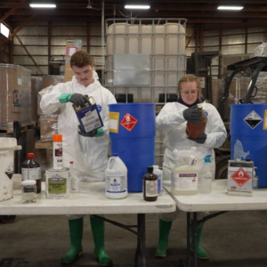 Two technicians in protective suits inspect and sort various household and chemical waste containers at folding tables inside a facility.