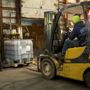 A forklift operator positioning a yellow Yale forklift near a large intermediate bulk container (IBC) tote on a floor scale in a warehouse.