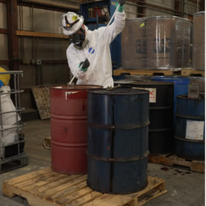 A technician wearing full PPE and a respirator uses a sampling tube on a chemical drum inside a warehouse.