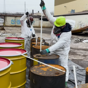 Two environmental technicians in protective hazmat suits and respirators taking liquid samples from chemical drums using glass coliwasa tubes.
