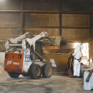 A Bobcat skid-steer loader dumping materials into a wooden hopper while a worker in a white hazmat suit stands nearby holding a shovel.