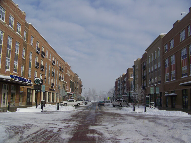 A quiet street scene in winter, featuring brick buildings on both sides and a snow-covered road. Vehicles are parked along the curb, and the sky is overcast with clouds, creating a muted atmosphere.