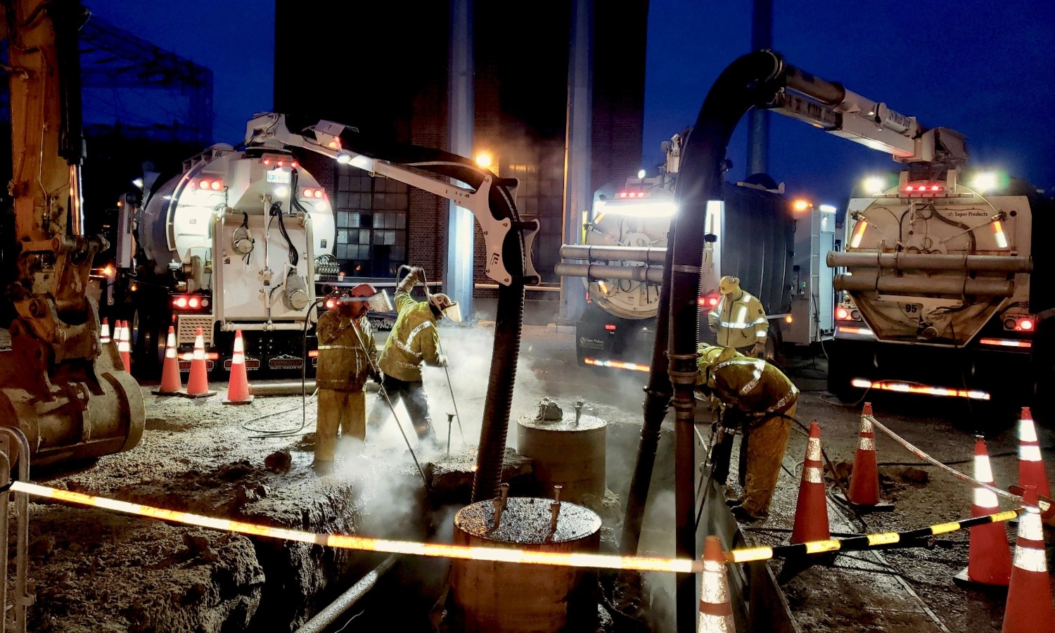 Home Nighttime hydro excavation operation with workers using pressurized lances and vacuum boom hoses around steam-emitting underground utility pipes.