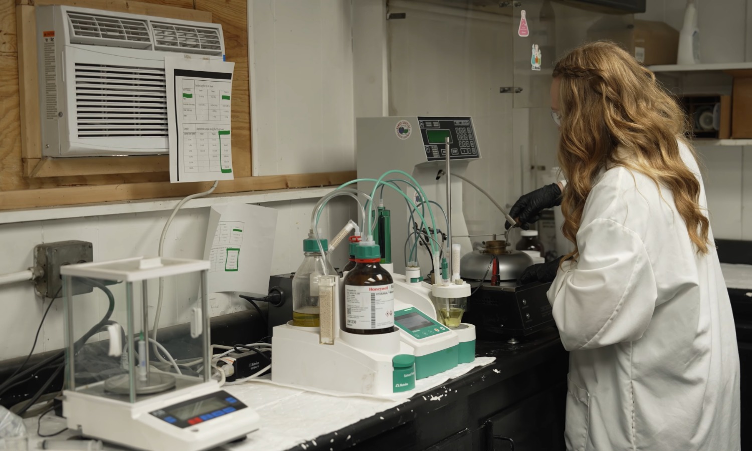 Home A laboratory scene showing a woman in a white lab coat using equipment on a counter. Various glass containers, a digital scale, and measuring instruments are arranged on the surface, with an air conditioning unit mounted on the wall.
