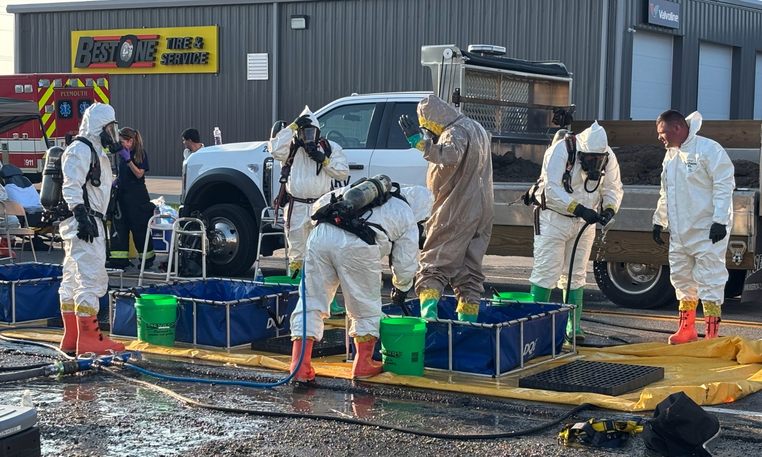 Home Hazmat team members in white protective suits and orange boots work at a decontamination site. They stand near blue containers filled with liquid, while a pickup truck and a fire truck are visible in the background.