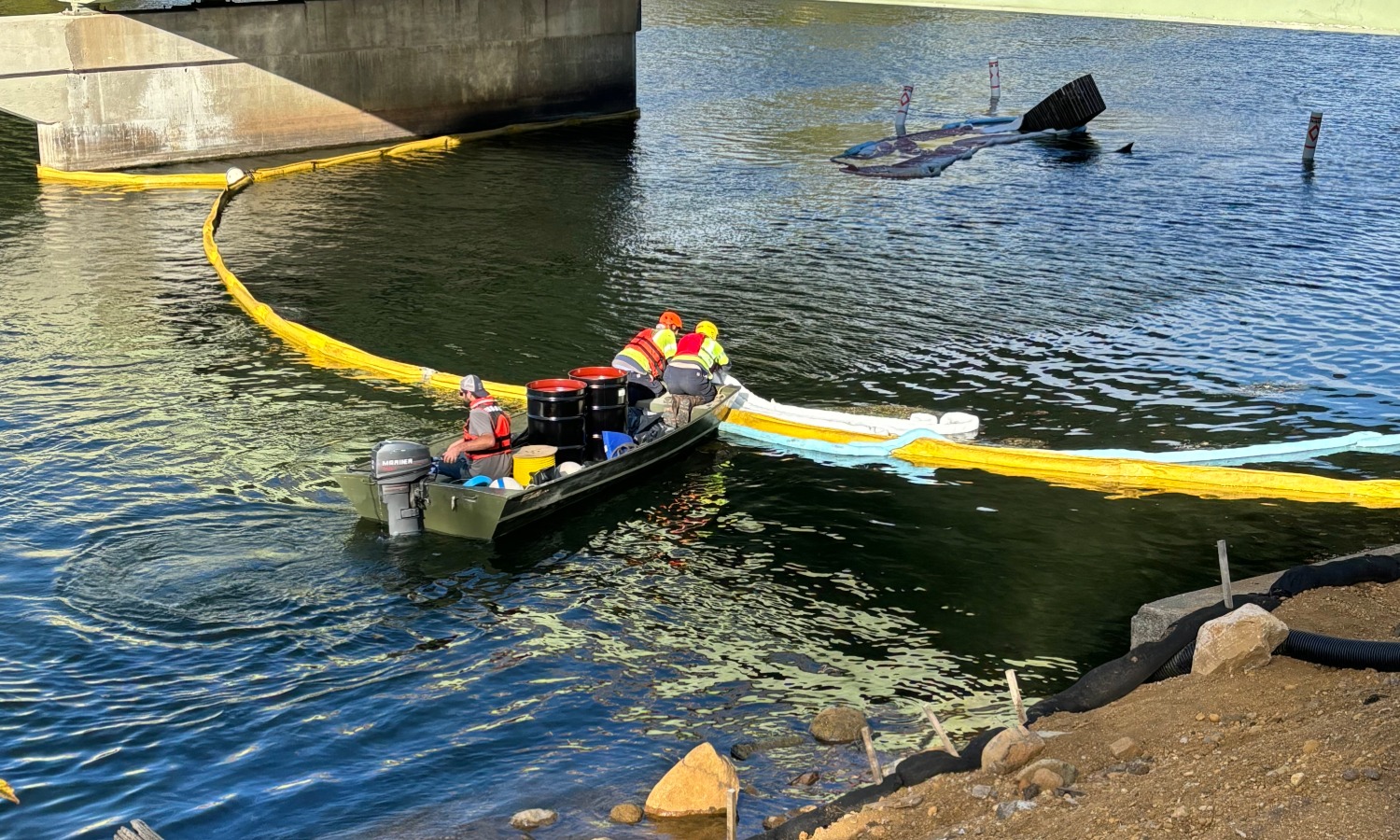 Home Two people in safety vests are in a small boat on a body of water, moving towards a floating containment barrier. The scene features a bridge in the background and a submerged object partially visible. Yellow and blue materials are in the water.