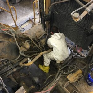 Industrial Services A person wearing a white coverall and yellow boots is seated inside a machinery compartment, working among numerous tangled hydraulic hoses and wires. The environment appears dusty, with tools and parts visible in a workshop setting.