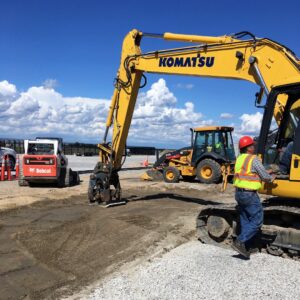 Industrial Services A construction site featuring three pieces of heavy equipment, including a yellow Komatsu excavator, a Bobcat loader in the background, and a backhoe near the center. A worker in a safety vest and hard hat operates the excavator on a clear day with blue skies.