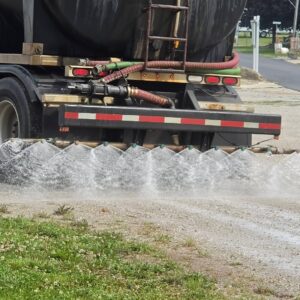 Seasonal Services A close-up of a water truck showing the rear spraying mechanism discharging a stream of water onto the ground. The truck has hoses attached, with a visible section of the road and grass in the foreground.