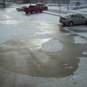 Seasonal Services A parking lot covered in a layer of melting ice and light snow, revealing patches of wet concrete. A tan car is parked on the right, while several vehicles, including red trucks, are visible in the background. This scene illustrates the effectiveness of products like ICE BAN DeIcer and ICE Ban Anti-Icer.