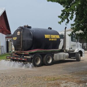 Seasonal Services A large black tank truck labeled "Dust Control" is spraying water onto a gravel road. In the background, there are white and red agricultural buildings and trees, indicating a rural setting.