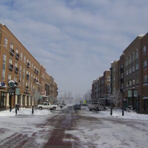 Seasonal Services A snow-covered street lined with brick buildings on both sides. Cars are parked along the road, and the scene features street signs and lampposts. The sky is cloudy, and a light layer of snow covers the ground.